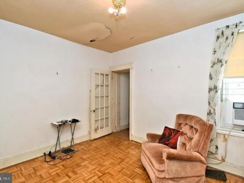 Empty room with hardwood floors, peeling paint ceiling, and a small brass chandelier. A single window with floral curtains and an air conditioner. A brown armchair with a pillow and a small table with electronics.