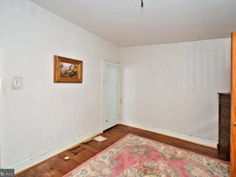 Empty room with white painted walls, hardwood floor, a pink floral rug, a framed landscape painting, and a dark wooden dresser in the corner.