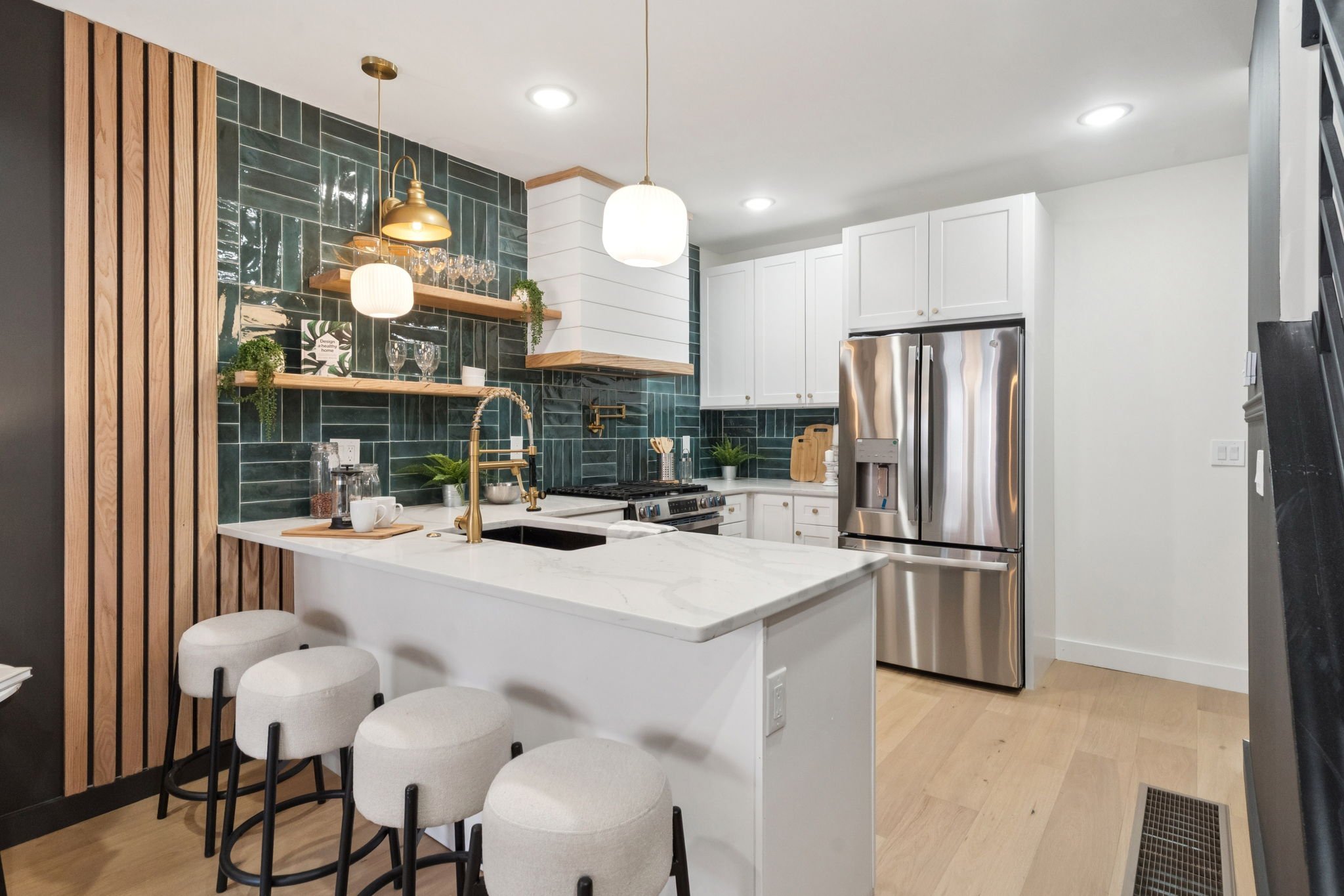 Modern kitchen with dark green tile backsplash, white cabinets, stainless steel refrigerator, and a built-in sink with gold fixtures. Open shelving displays glassware and plants. There is a white marble island with three white barstools, hanging pend