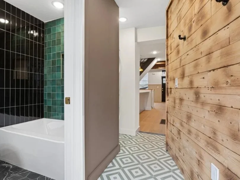 View of a hallway leading to a kitchen with wood flooring, a part of a bathroom with black and green tiles, and a wooden accent wall.