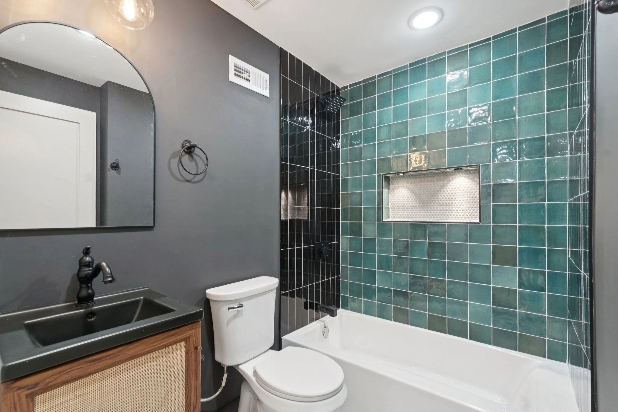 A modern bathroom featuring a dark gray wall with a mirror, a black sink with a matte black faucet, a white toilet, and a bathtub with green tiles. The shower area has black and dark green tiles with a recessed shelf and a black showerhead.