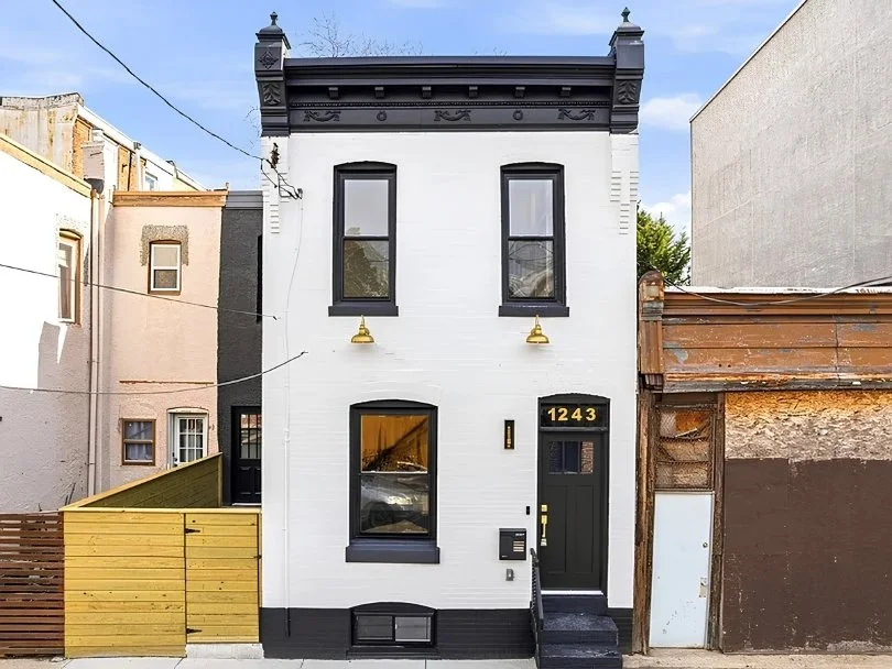 A narrow, three-story white brick townhouse with black trim and a black door numbered 1243, flanked by neighboring buildings, one with a yellow fence.