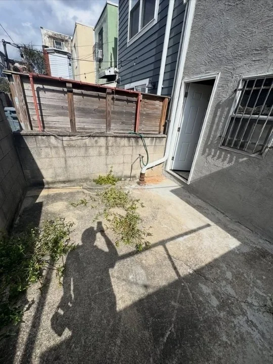 Small backyard with concrete floor, overgrown weeds, wooden fence, and house wall with window and door.