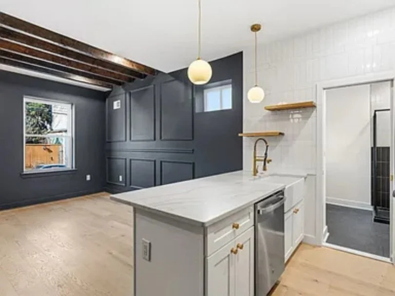 Modern kitchen with white cabinets, marble countertop, gold fixtures, and a dark wall with wooden beams ceiling.