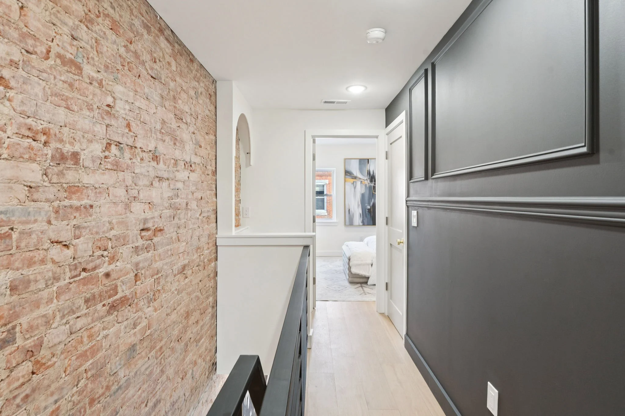 Hallway with exposed brick wall on the left, gray and white walls, and a glimpse into a living area with a sofa and artwork in the background.