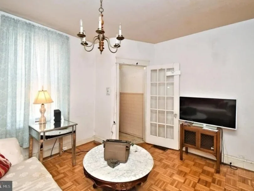 Living room with white walls, wood flooring, a window with light blue curtains, a chandelier hanging from the ceiling, a white couch with a red pillow, a small glass-top side table with a lamp and digital device, a round marble coffee table with a ba