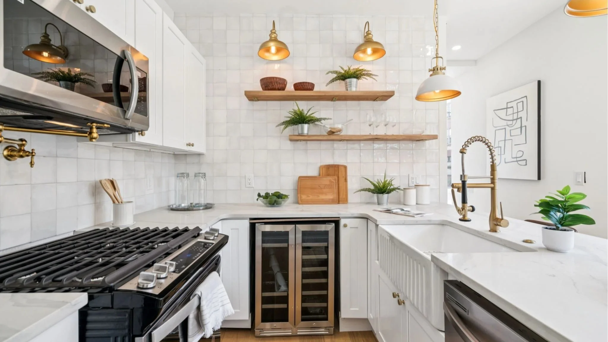 Modern kitchen with white cabinets, marble countertops, and gold fixtures. Open shelving with plants and bowls, gas stove, wine cooler, and a sink with a gold faucet. Decor includes framed artwork and small plants.