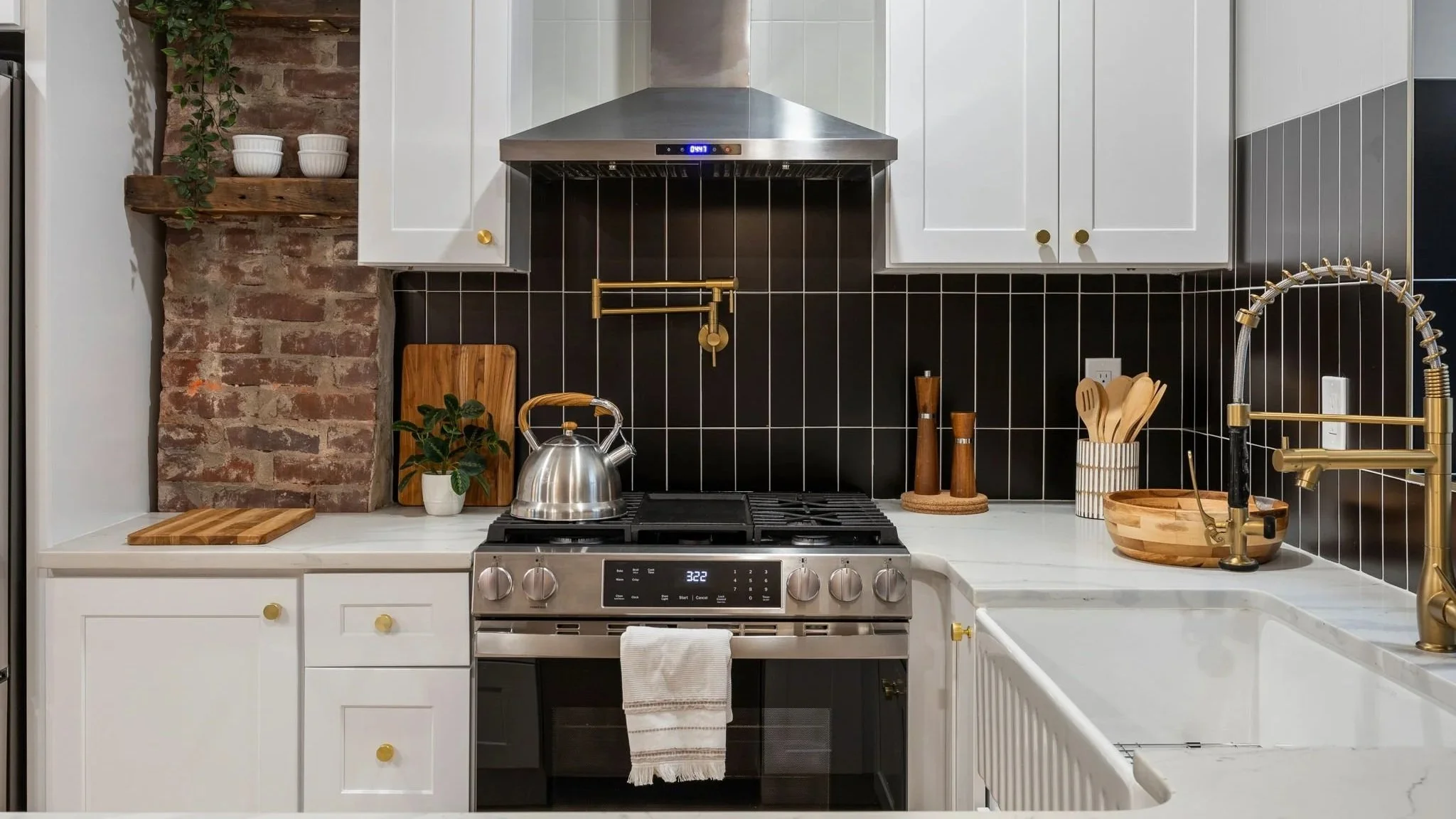Modern kitchen with white cabinets, black tiled backsplash, stainless steel oven, brass faucet, teapot, cutting boards, plant, and decorative bowls.