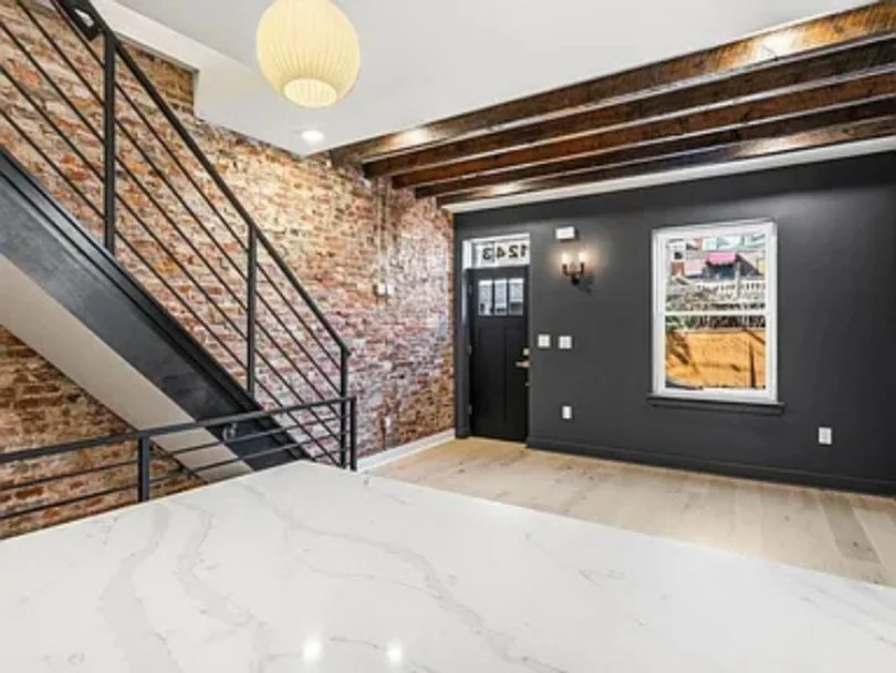 Interior of a modern apartment with exposed brick wall, black door, large window, dark gray wall, wood beams on ceiling, and a staircase with black metal railing.