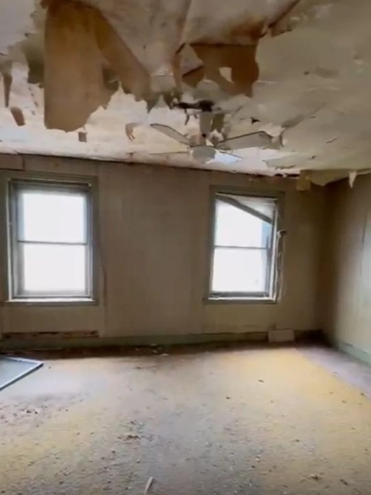 Empty room with two windows and a damaged ceiling with peeling paint and debris.