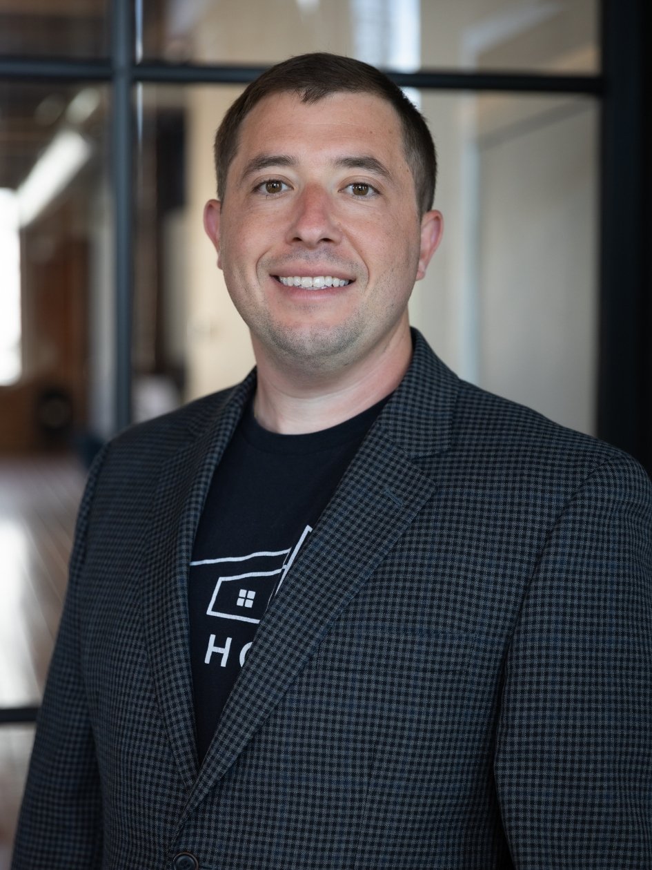 A smiling man with short dark hair wearing a black shirt and a black and gray checkered blazer, standing indoors with a blurred office background.