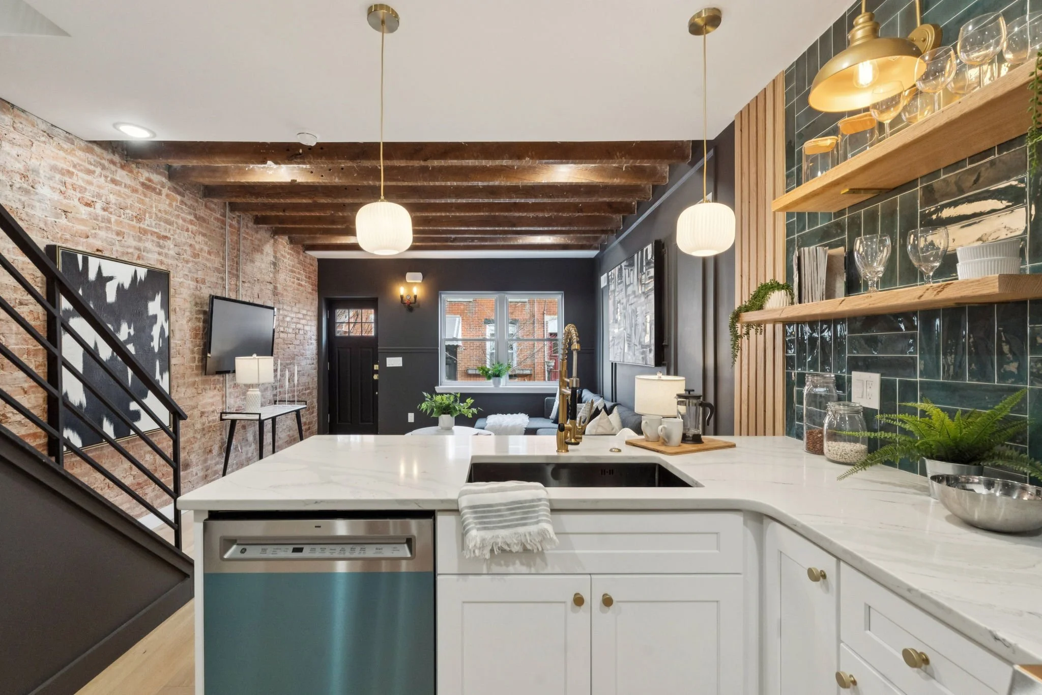 A modern kitchen with a white marble island, gold hardware, and open wooden shelves with glassware. The ceiling has wooden beams, and there is a living room with a dark wall, a window, and a black door in the background.