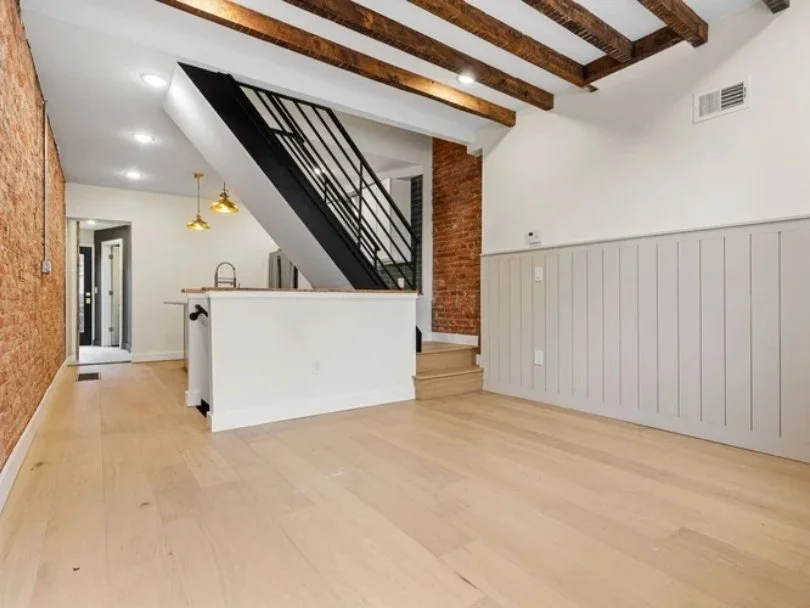 Empty living room with wooden floors, exposed ceiling beams, white walls, and a staircase with black metal railing leading upstairs.