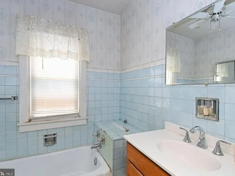 A vintage bathroom with a white bathtub, light blue tiled walls, and a small window with lace curtains. There is a washing machine next to the tub and a wooden vanity with a white sink and silver fixtures. A large mirror is above the sink.