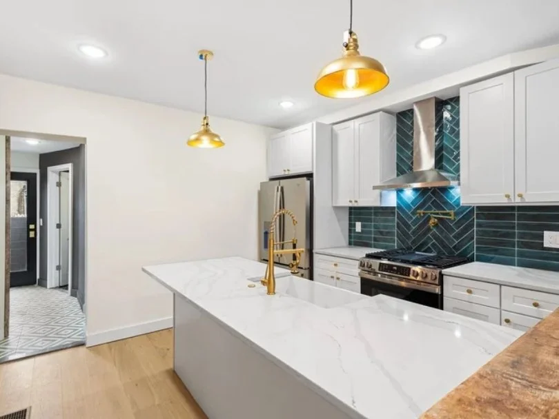 Modern kitchen with white cabinets, a marble island, and blue tile backsplash. Features gold accents including a faucet and pendant lights, with stainless steel appliances.