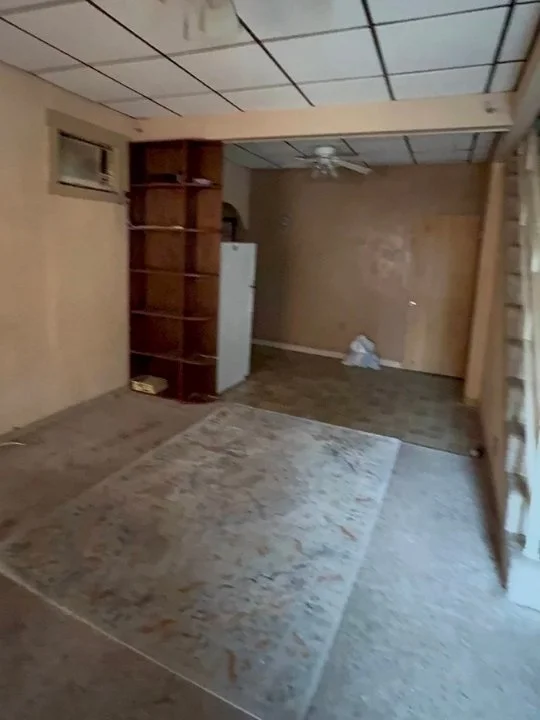 Empty room with wooden walls, a ceiling fan, and a wooden bookshelf near a white refrigerator. The floor has a large, worn rug, and there are some items on the floor in the back corner.