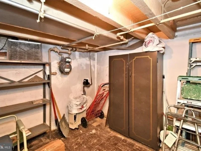 Basement storage area with a metal shelving unit, a garden cart, a large cabinet, and various household items, including a snow shovel, a fan, and a bag on top of a small appliance.