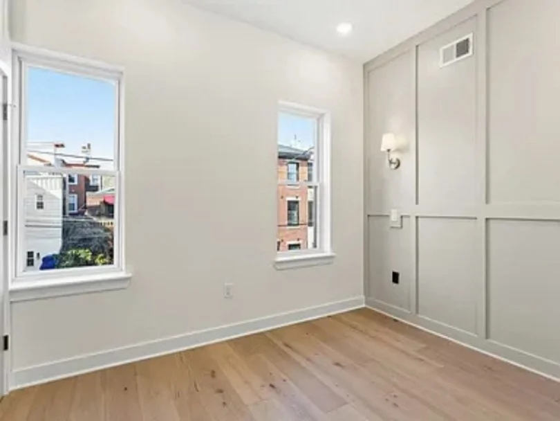 Empty room with two windows overlooking neighboring buildings, white walls, light wood floors, and wall-mounted light fixture.