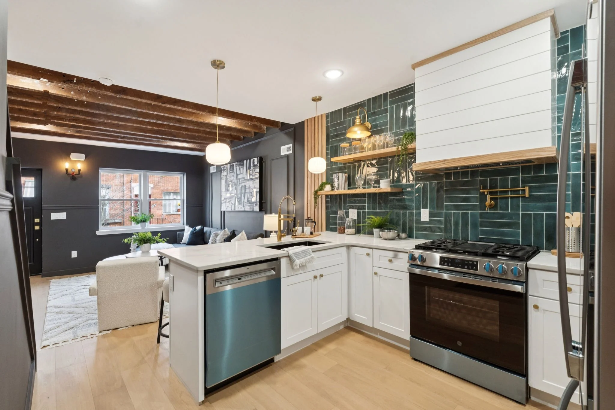 A modern kitchen with white cabinets, a blue island, a green tiled backsplash, and wood accents. Open shelving displays glassware and plants. In the background, a living area has a dark wall, large window, and ceiling beams.