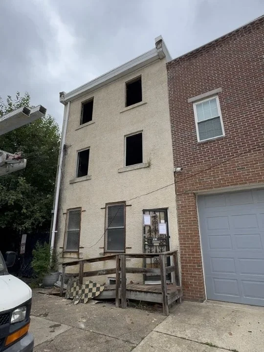 A four-story building with a worn, beige exterior, missing windows on the upper floors, boarded-up lower windows, and a small, accessible front porch with a simple railing, attached to a brick building with a garage door.