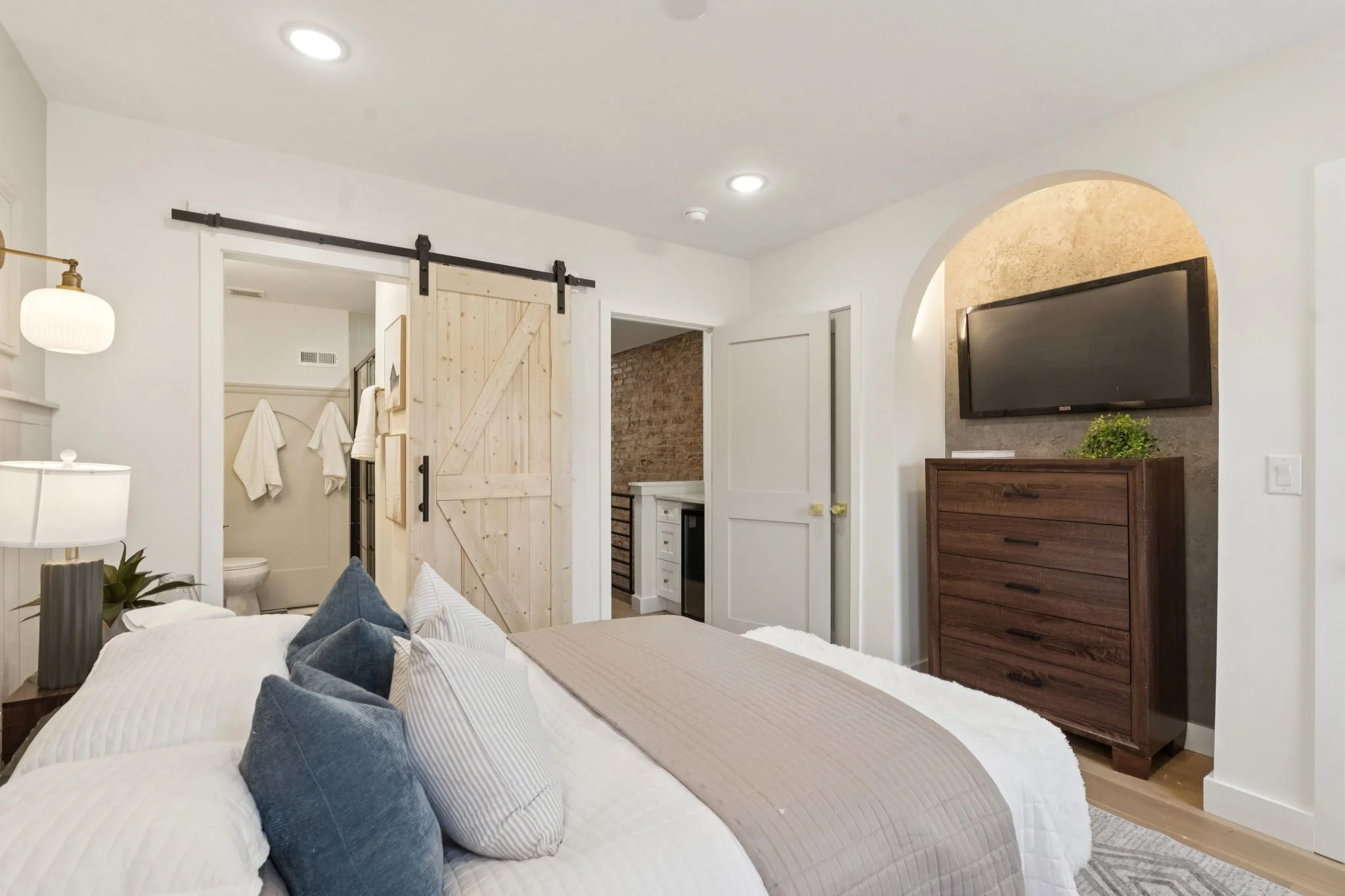 Interior of a bedroom with a white bed, blue pillows, and a beige blanket. To the left, there's a note lamp and plant on a small table. In the background, there's a sliding barn door on a black track, leading to a bathroom with towels and a toilet. T