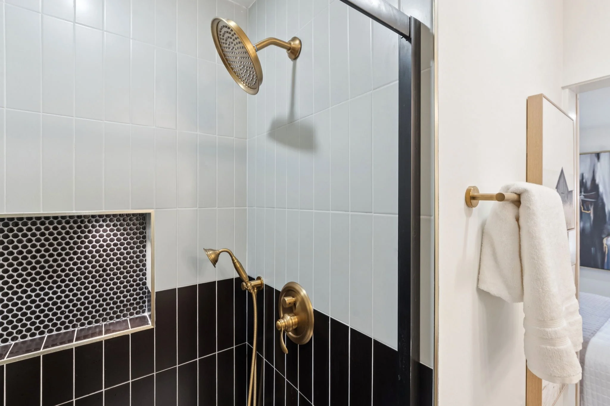 A bathroom shower with white tiled wall, black tiled lower section, gold fixtures, and a towel hanging on a gold towel bar.