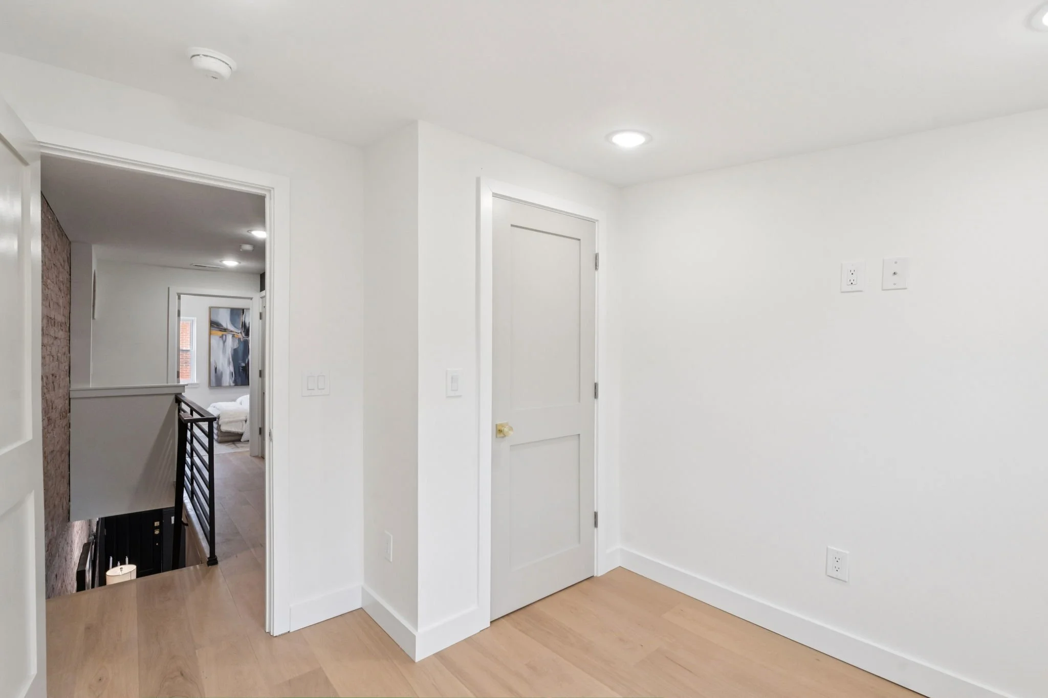 A bright, minimalist interior space with white walls, a closed white door, and wood flooring. Part of a hallway visible with additional rooms and artwork in the background.
