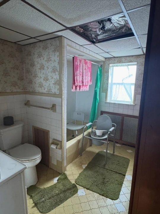 An old, cluttered bathroom with peeling wallpaper, a toilet, a small shower area with a pink towel hanging and a pillow, a window with broken and taped glass, and two green bath mats on a yellow patterned floor.