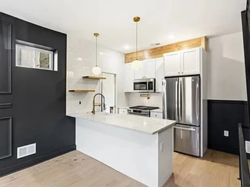 Modern kitchen with white cabinets, a stainless steel refrigerator, microwave, and oven. Black wall with decorative paneling, light wood flooring, and hanging pendant lights over white countertop island.