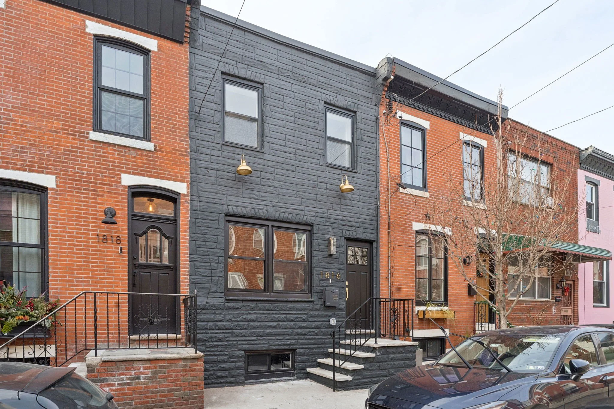 Row of brick and stone townhouses with front porches, stairs, and minimal landscaping on a city street, with parked cars in foreground.