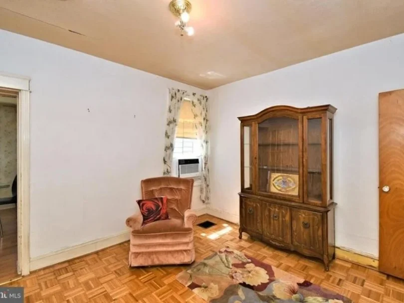 Empty living room with wood floor, white walls, single window with floral curtain, pink armchair with throw pillow, wooden china cabinet, small rug, ceiling light fixture, and door on the right.