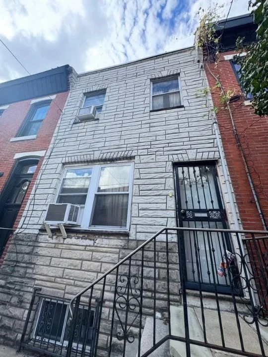 Front view of a narrow, three-story row house with a white brick facade and a black iron security door, railing, and window air conditioning unit, with neighboring brick row houses and a partly cloudy sky.