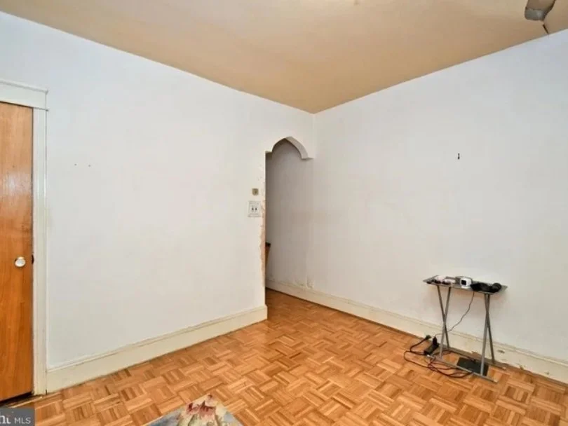 Empty room with white walls, a wooden floor, a small table with electronic items, and an open doorway with an arched top.