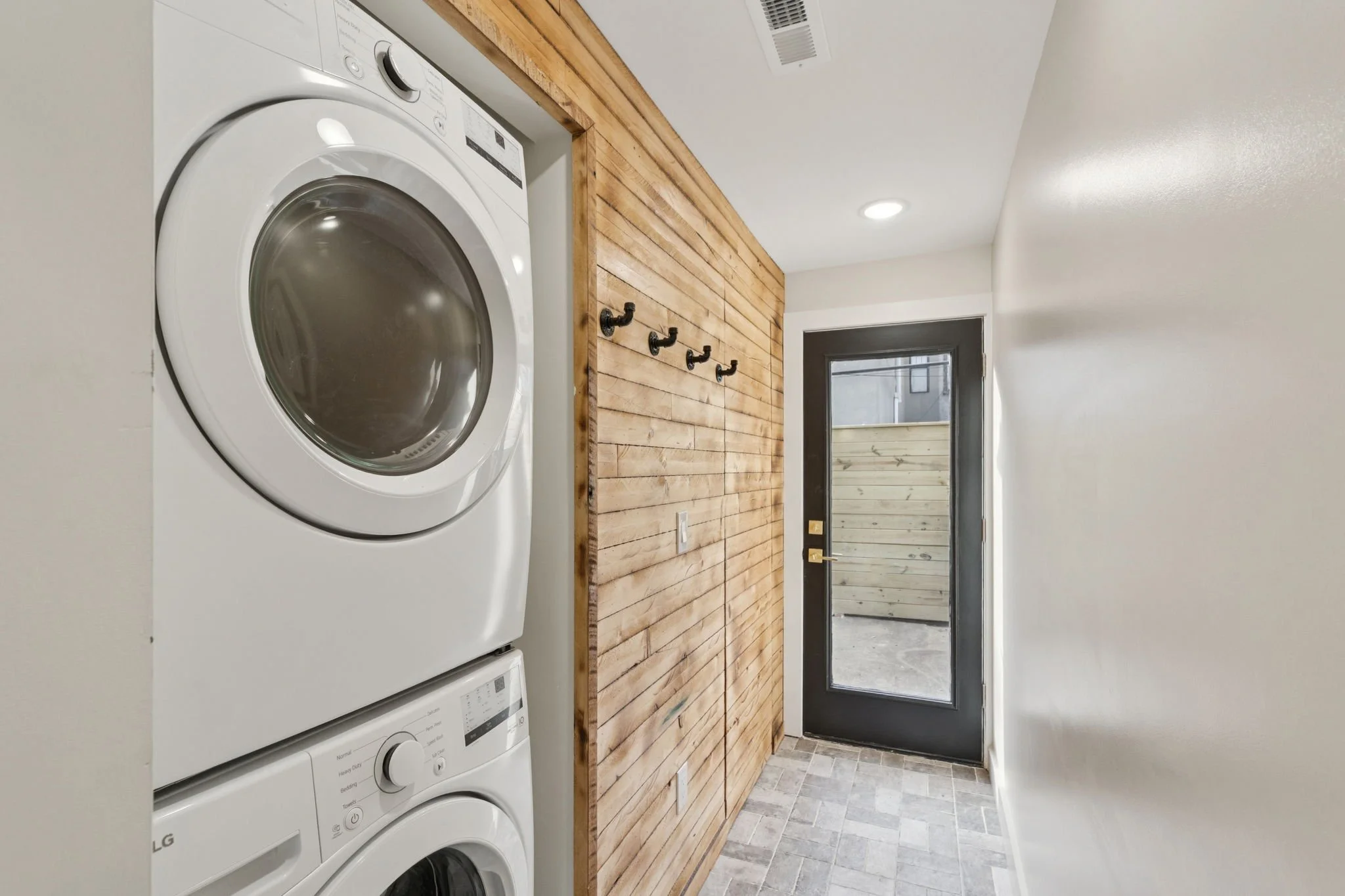 A laundry room with stacked front-load washer and dryer, a wooden slat accent wall, hooks on the wall, and a door leading outside.