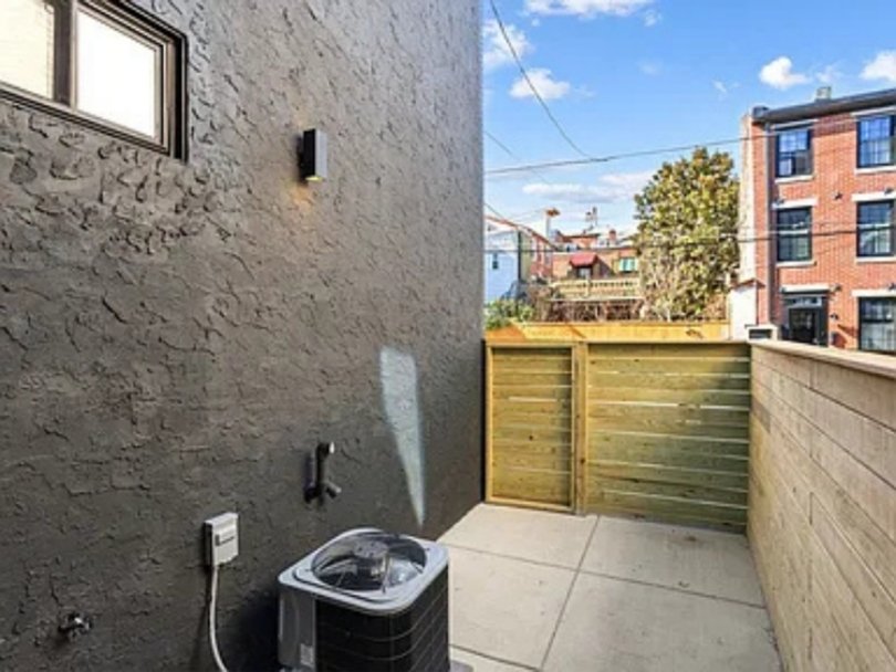 Small outdoor alley or patio area with a washer, privacy fence, and neighboring brick buildings in the background during daytime.