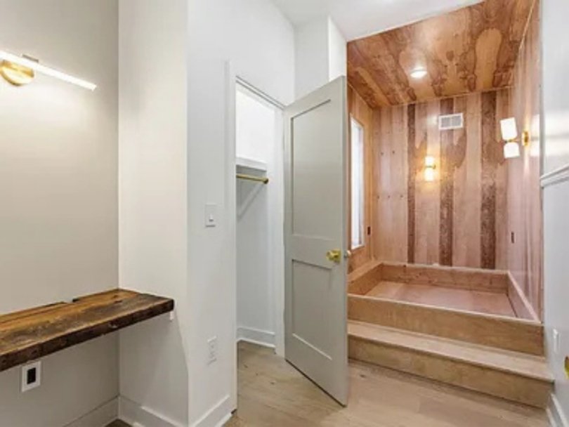 Interior of a room with white painted walls, a wooden countertop on the left, an open white door leading to a small closet, and a wood-paneled alcove with wall sconces on the right. The floor is light-colored wood.