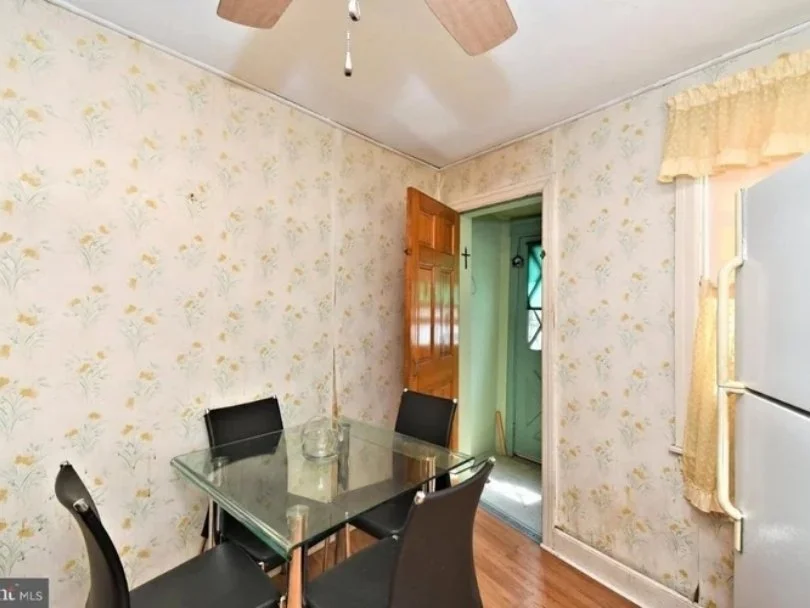 A dining area with a glass-top table and four black chairs, floral wallpaper, a ceiling fan, a refrigerator, a window with a yellow valance, and a door leading to the outside.