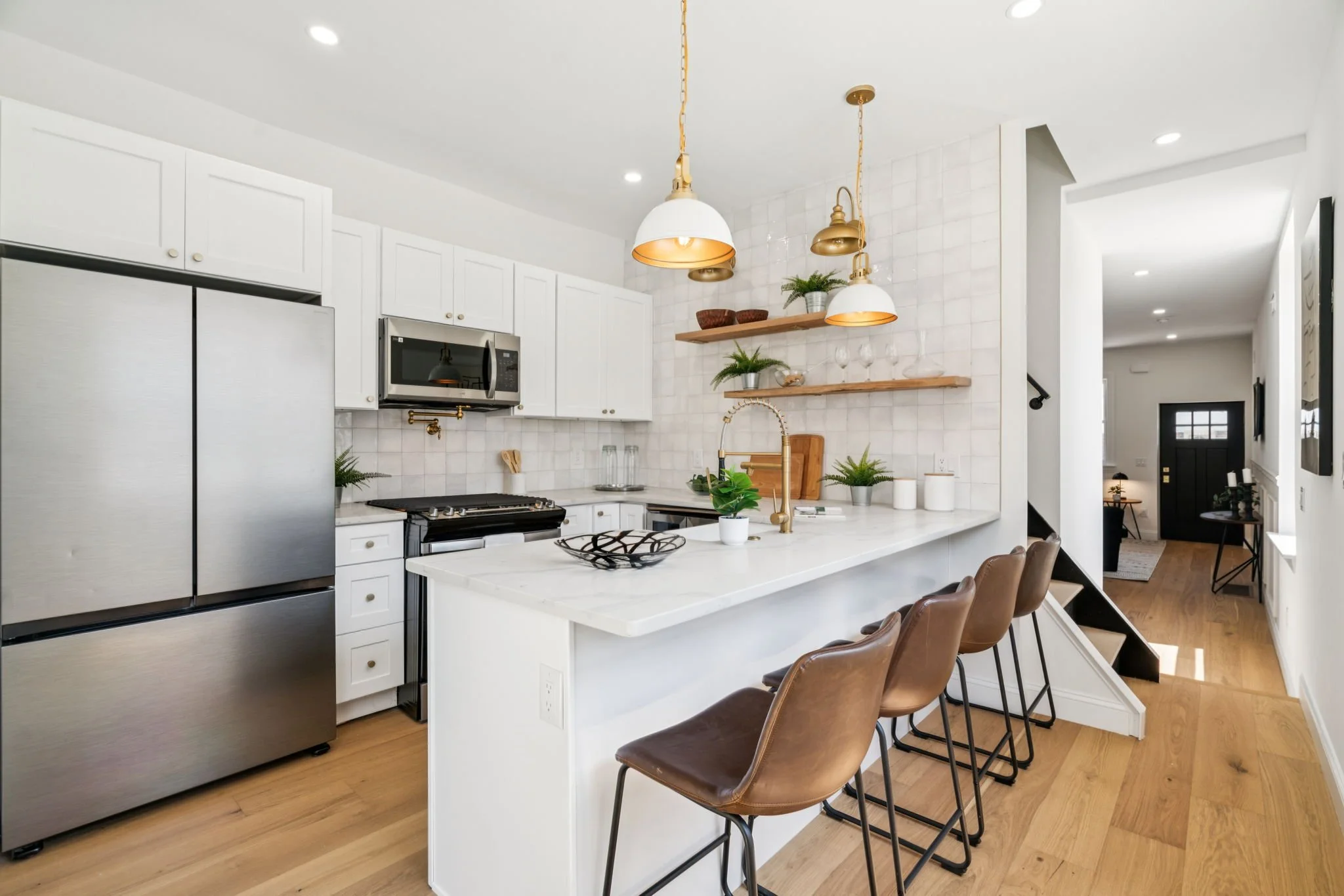 Modern kitchen with white cabinets, stainless steel refrigerator, microwave, and a white island with a sink. Three brown chairs are placed at the island. There are two wooden shelves with plants, bowls, and glasses. Pendant lights hang above the island. Wood flooring and a hallway leading to other rooms are visible.