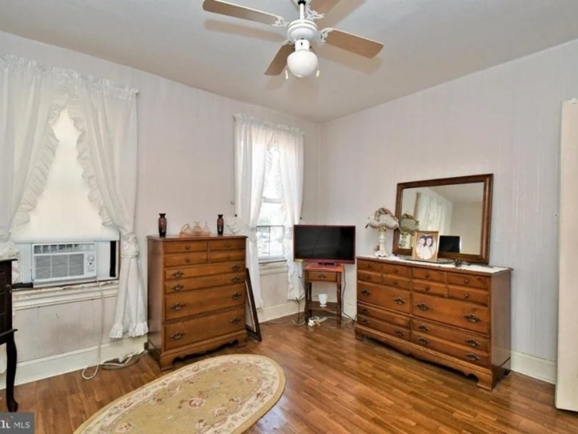 A bedroom with light-colored walls, hardwood floors, and white curtains. There are two wooden dressers, a small TV on a stand, a mirror, and decorative items on the furniture. An air conditioning unit is installed in the window.