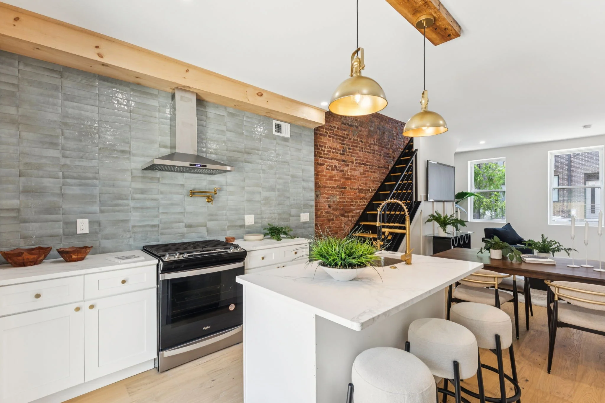 Modern kitchen with white cabinets, gray tile backsplash, stainless steel range hood, and an island with a gold faucet and plants. Adjacent open living space with large windows, brick wall, and a staircase with black railings.