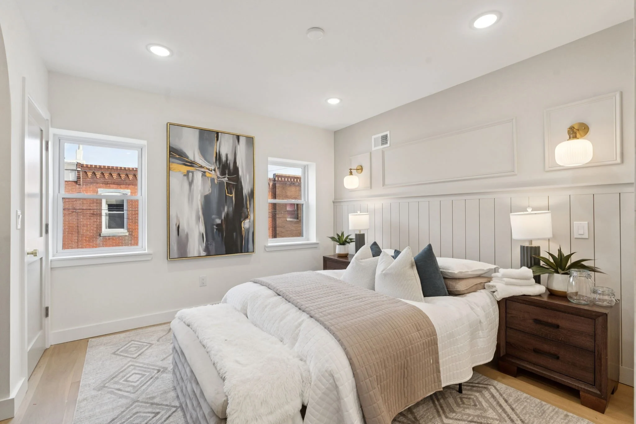 A bright bedroom with white walls and ceiling, featuring two windows, wall-mounted light fixtures, a large abstract artwork, a bed with multiple pillows and a beige bedspread, a dark wooden nightstand with a lamp, a small plant, and decorative jars o