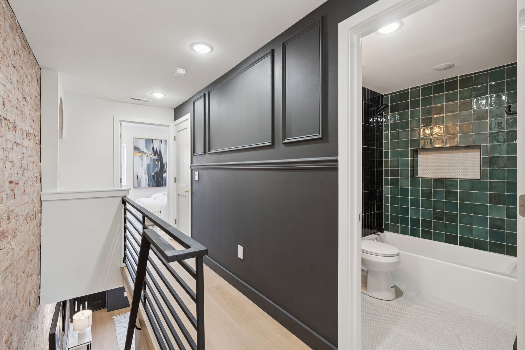 Interior view of a modern home bathroom with dark green tiled shower area, toilet, gray wainscoting on walls, white ceiling with recessed lighting, and a nearby hallway with a brick accent wall and contemporary artwork.