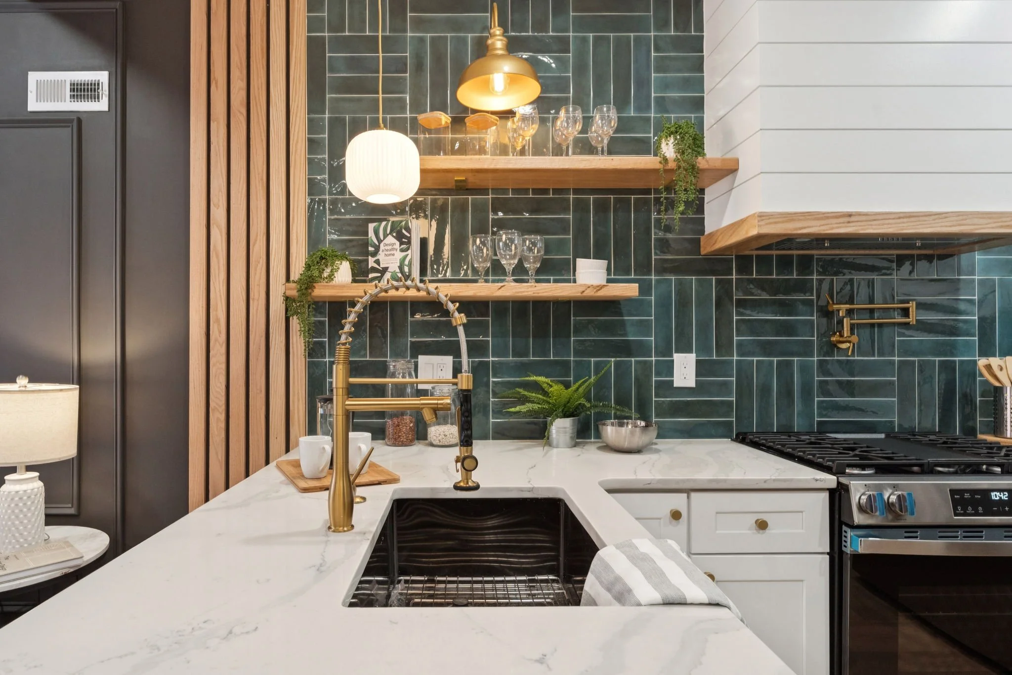 Modern kitchen with white marble countertop, gold faucet, and dark green tiled backsplash. Open wooden shelves hold glassware and small potted plants. Black and white cabinets and a stovetop visible.