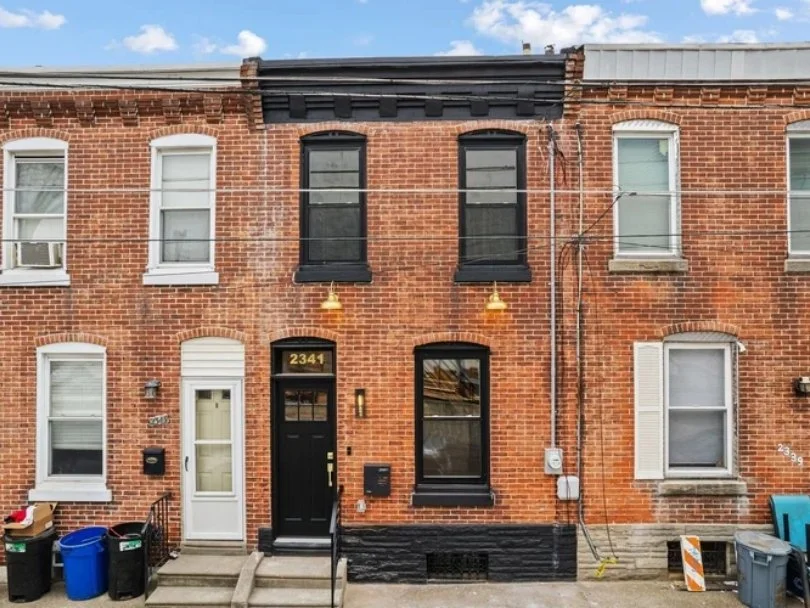 A brick rowhouse with black and white window shutters and an entrance with house number 2341, featuring a door and window on the front brick facade, two black pendant lights hanging, and neighboring houses with different window styles.
