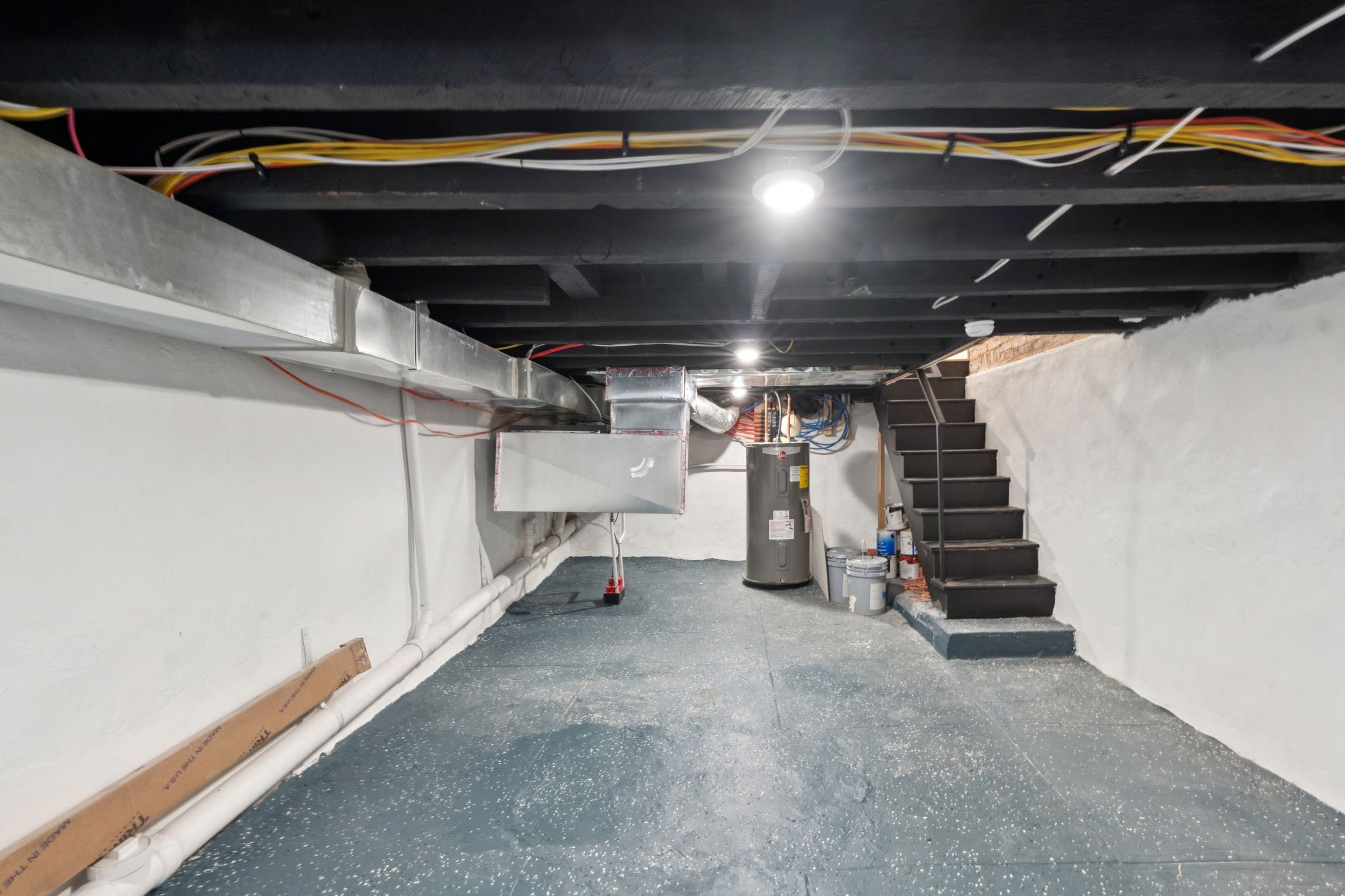 Underground basement with exposed ceiling, electrical wiring, ductwork, water heater, pipes, black stairs, and construction supplies.