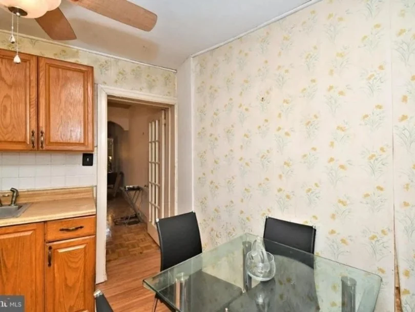 A dining area with a glass table and two black chairs, adjacent to a kitchen with wooden cabinets and a ceiling fan, and a doorway leading to another room.