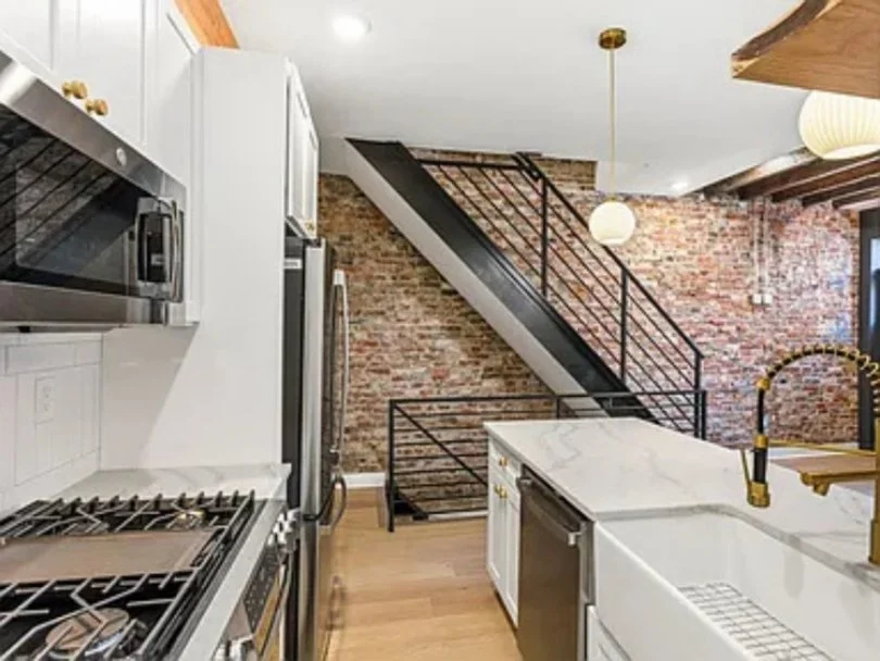 Modern kitchen with white cabinets, marble countertops, stainless steel appliances, and an exposed brick wall with a black metal staircase.