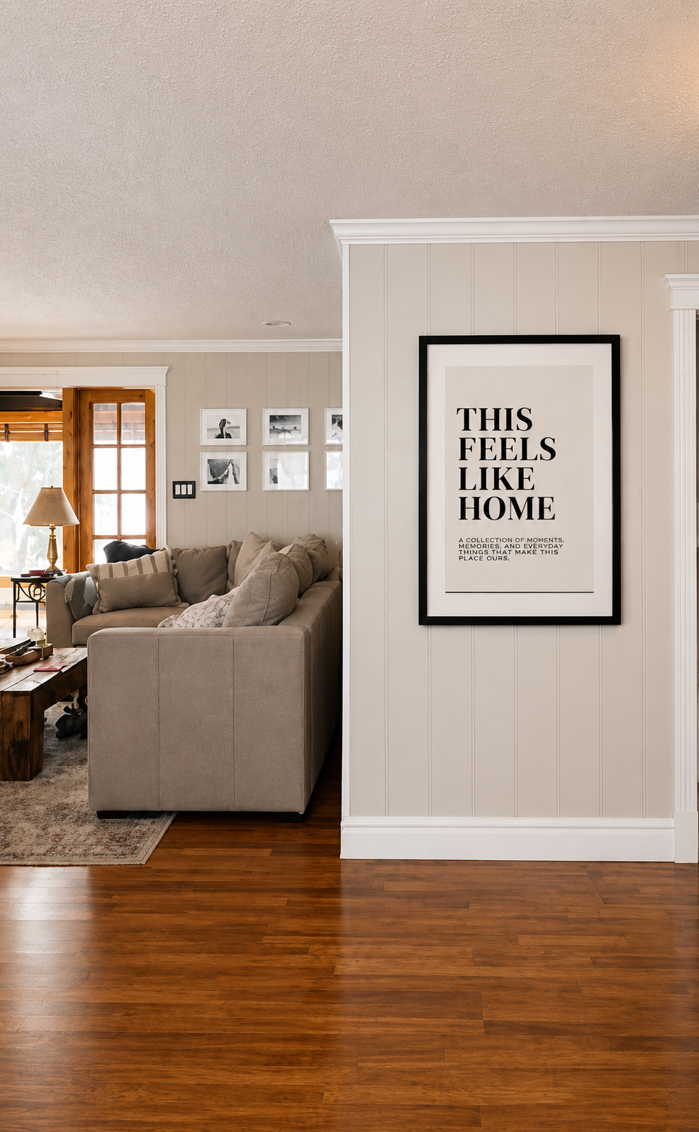 Living room with beige couch, wooden coffee table, and black and white photos on the wall. A framed print with the words 'This Feels Like Home' hangs on a white paneled wall. Hardwood flooring and a door leading outside are also visible.