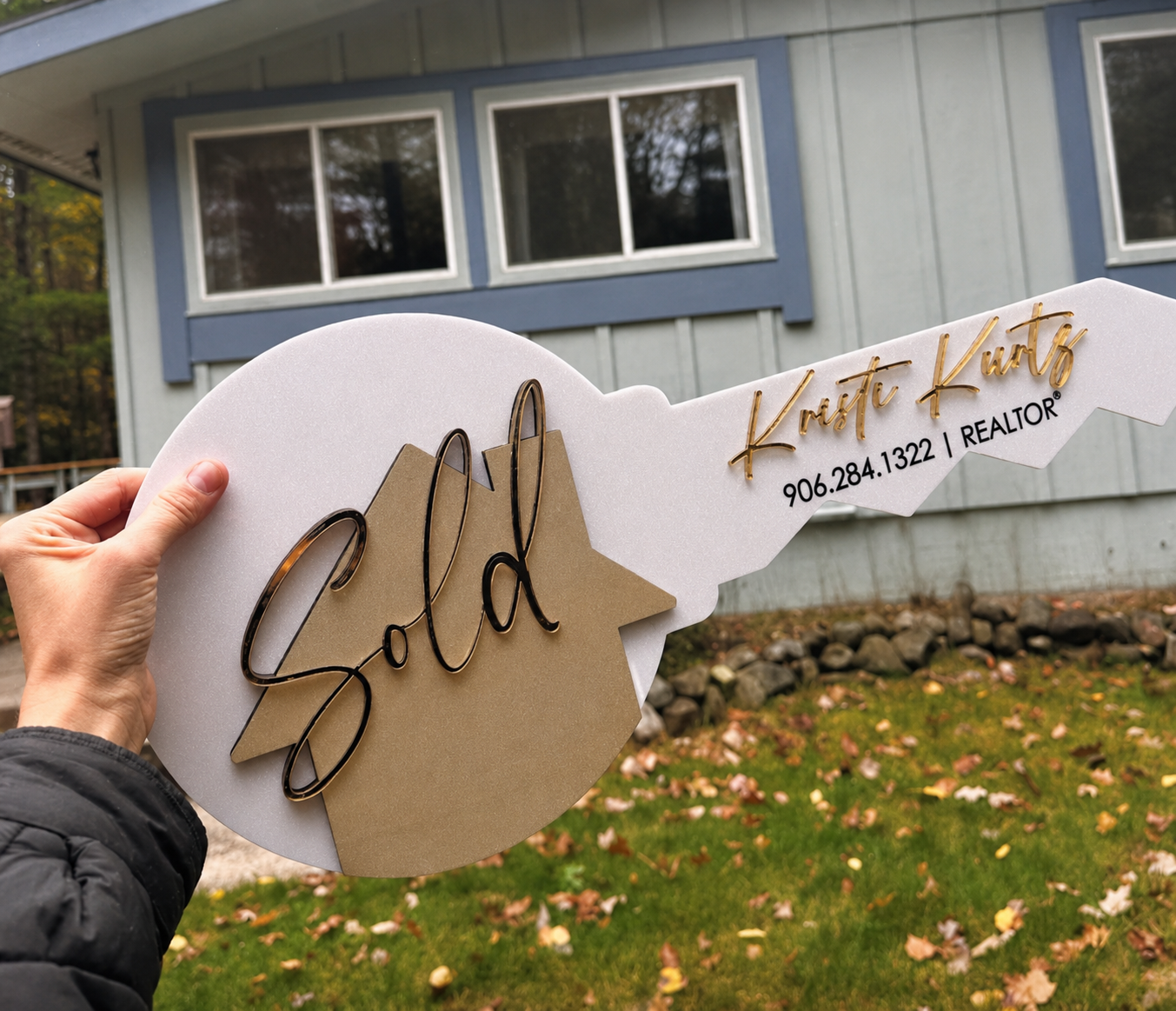 Person holding a large key-shaped sign with the word 'Sold' and the realtor's name, Krista Kurtz, along with phone number, in front of a house with blue trim and three windows.