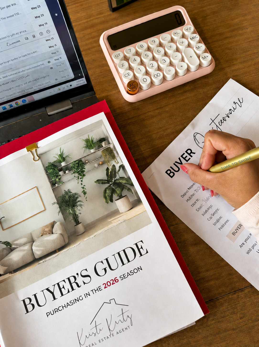 A person writing on a house buyer's guide with a pencil, placed on a wooden table, alongside a pink calculator, an open laptop, and real estate brochures with indoor plants on the cover.